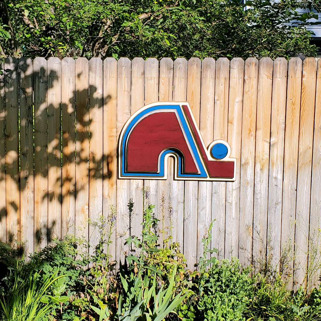 Wooden Nordique logo in 3D, painted in red, blue, and white on a wooden fence surrounded by greenery