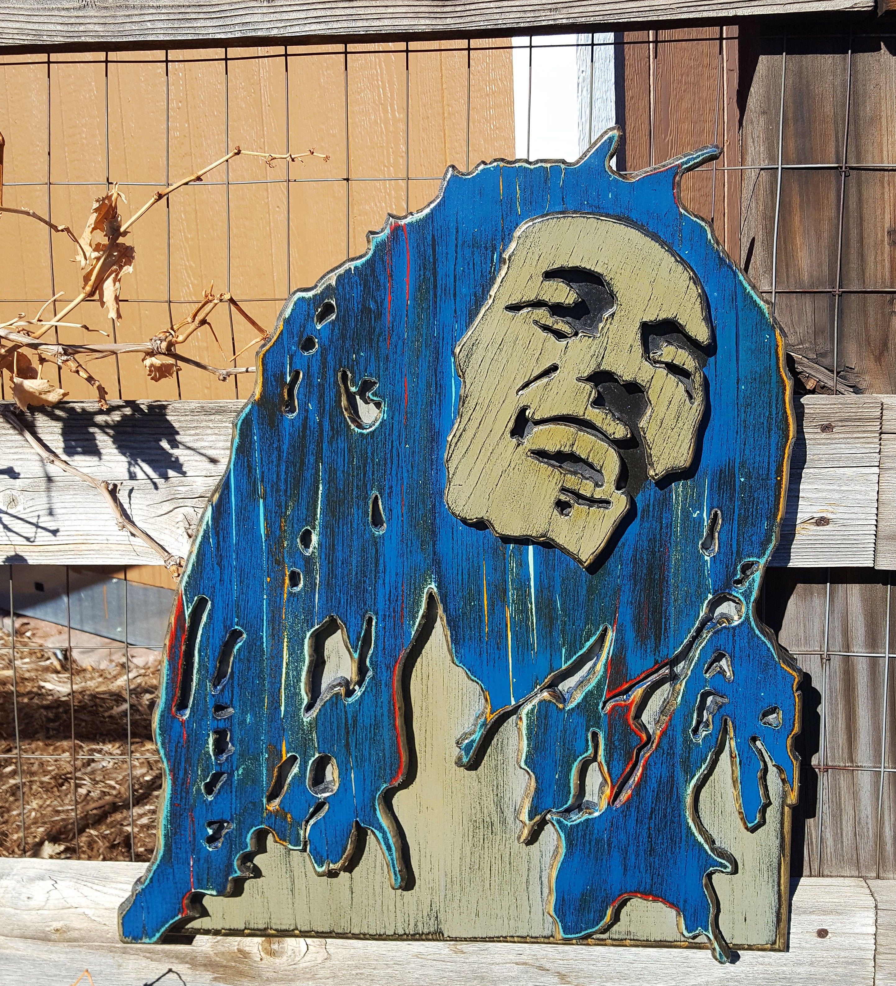 Wooden sculpture of Bob Marley's face and hair in blue and brown tones on a wooden background