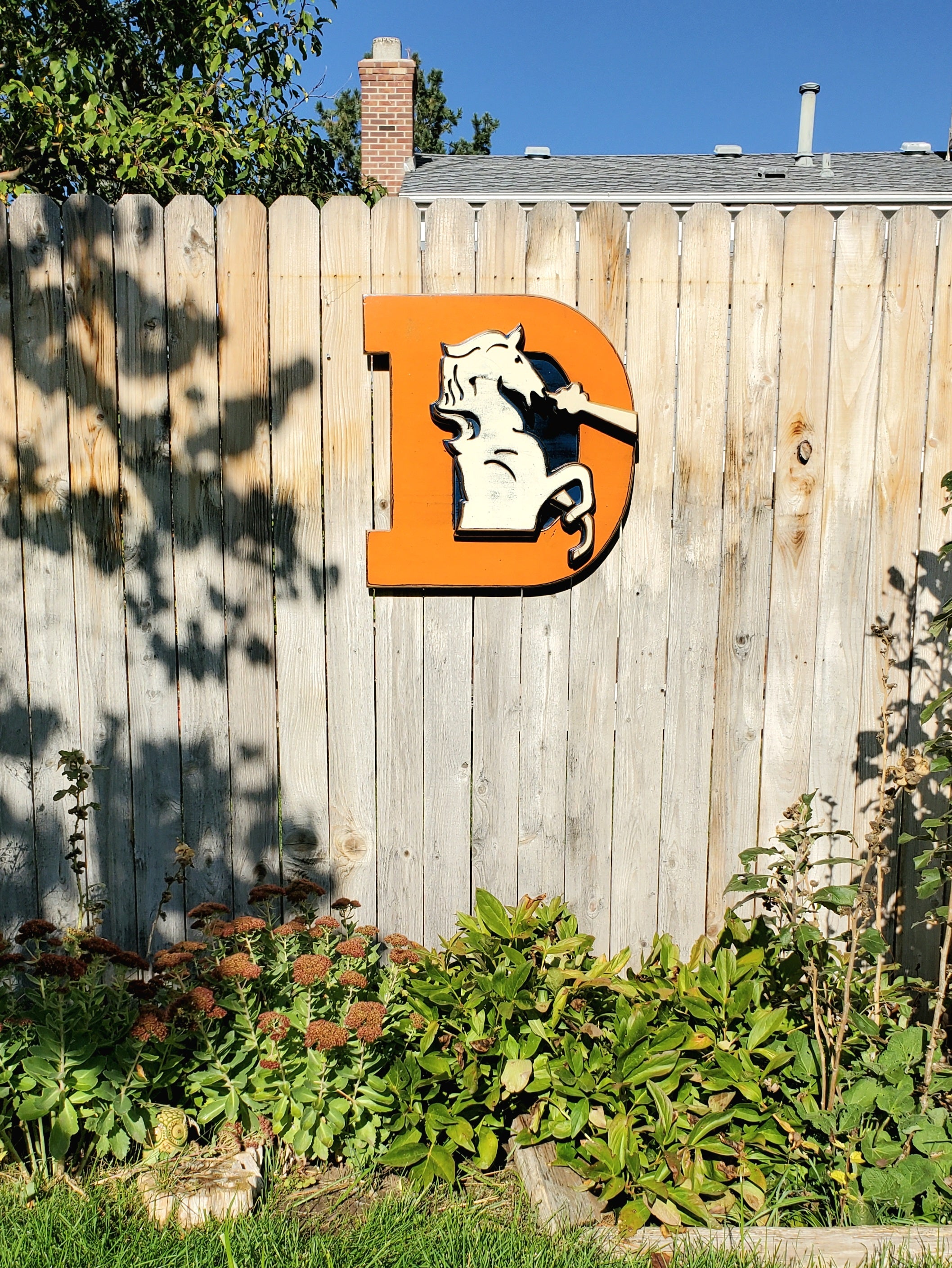 Wooden Broncos Retro "D" logo displayed on a wooden fence surrounded by trees and greenery 