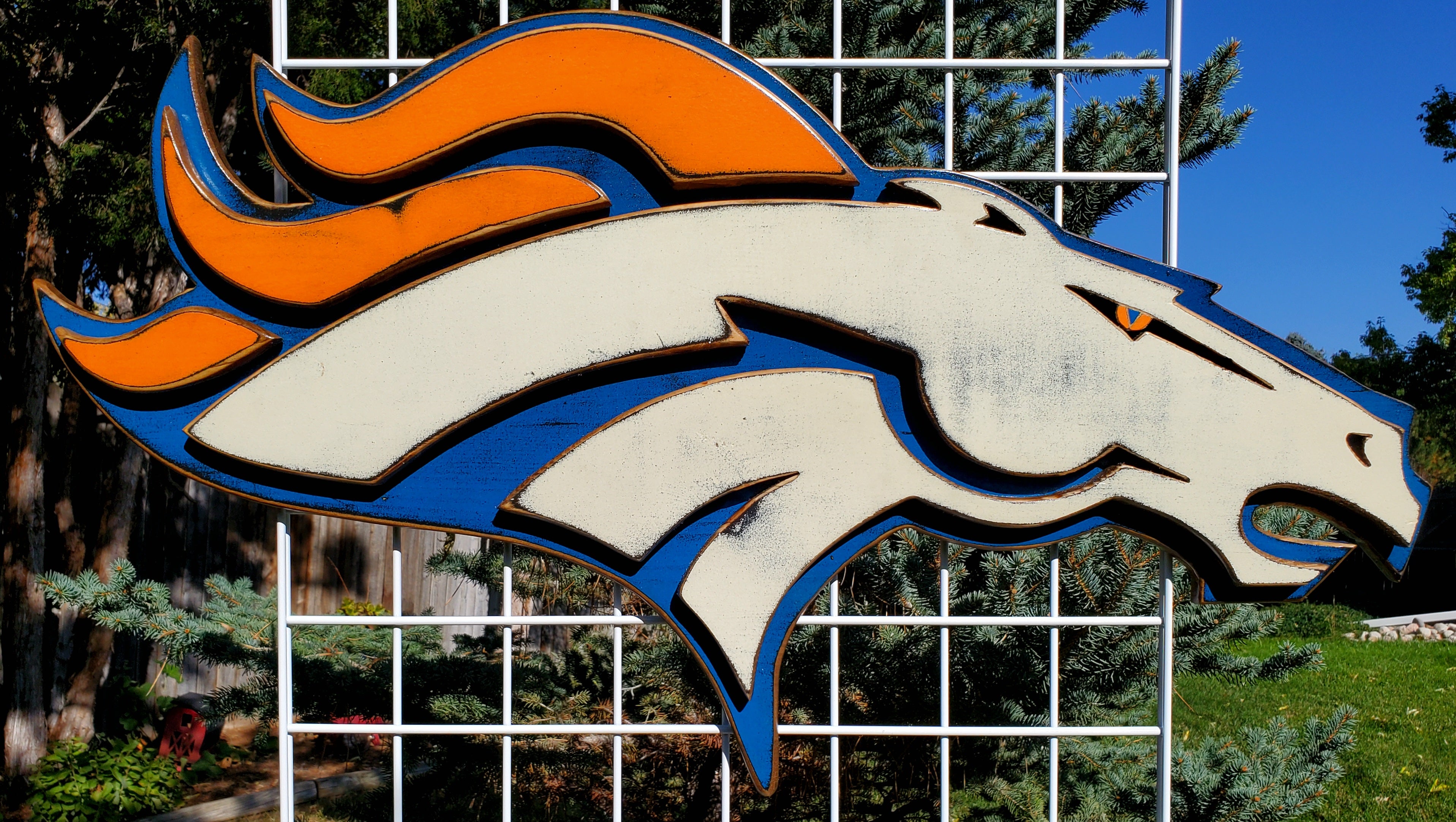 Denver Broncos modern logo on a metal grid with trees and greenery in the background