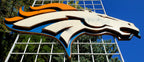 Denver Broncos modern logo displayed on a metal grid with trees and blue sky in the background