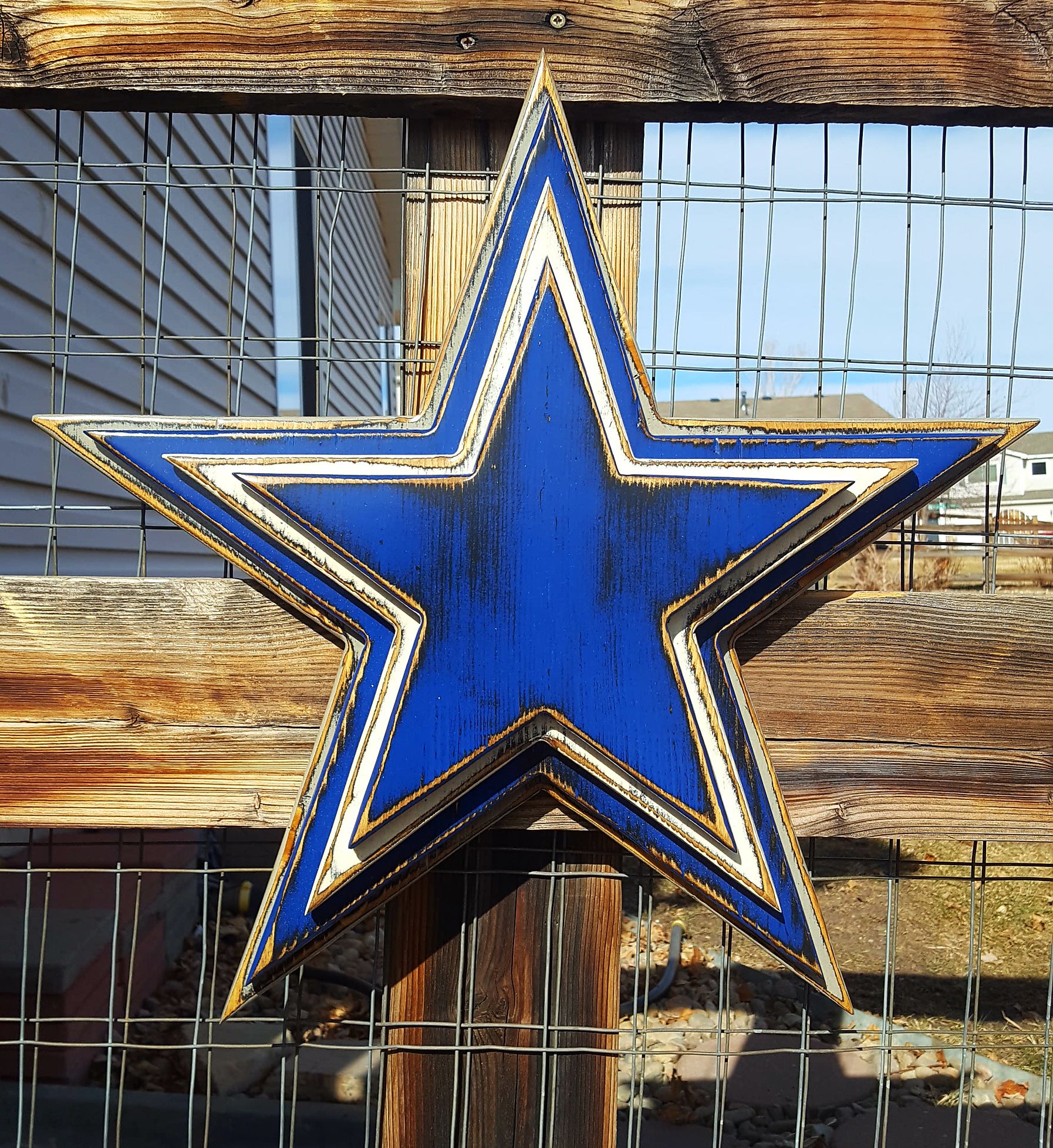 Cowboys star logo with four wooden layers alternating blue, white, blue, gray and displayed on a wooden fence.