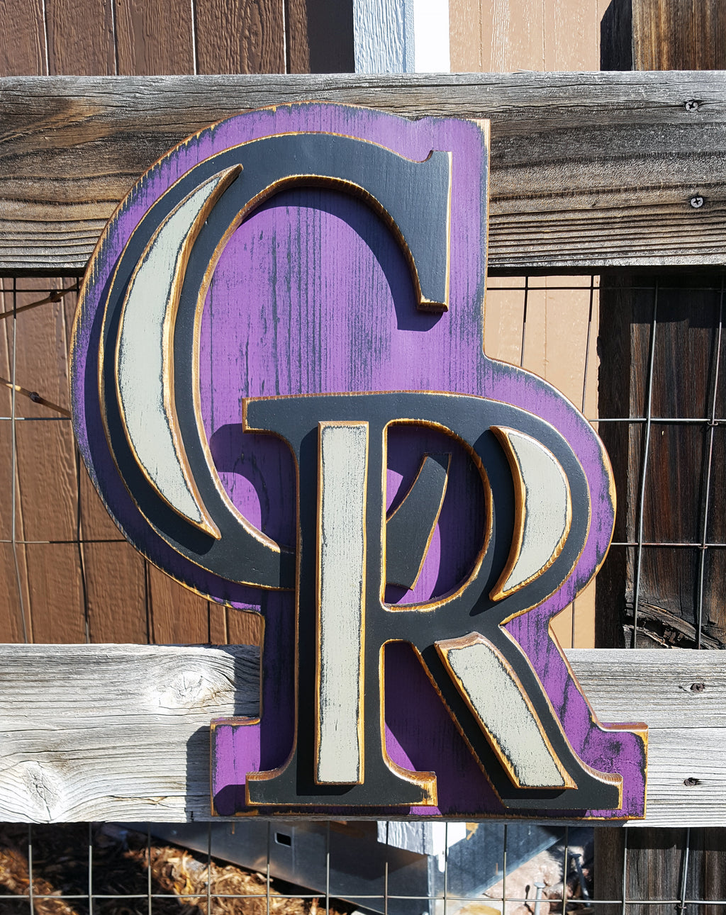 Decorative sign with Colorado Rockies "CR" logo with a purple background on a chicken wire fence